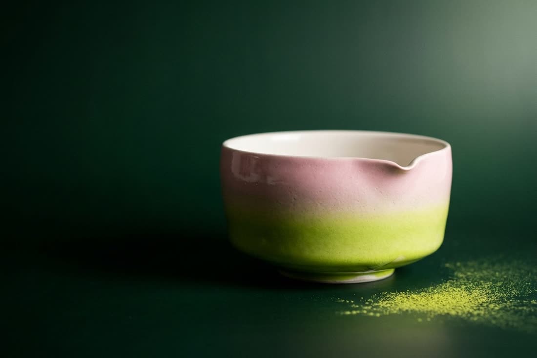 A pink and green glazed matcha bowl with a pouring spout beside scattered matcha powder