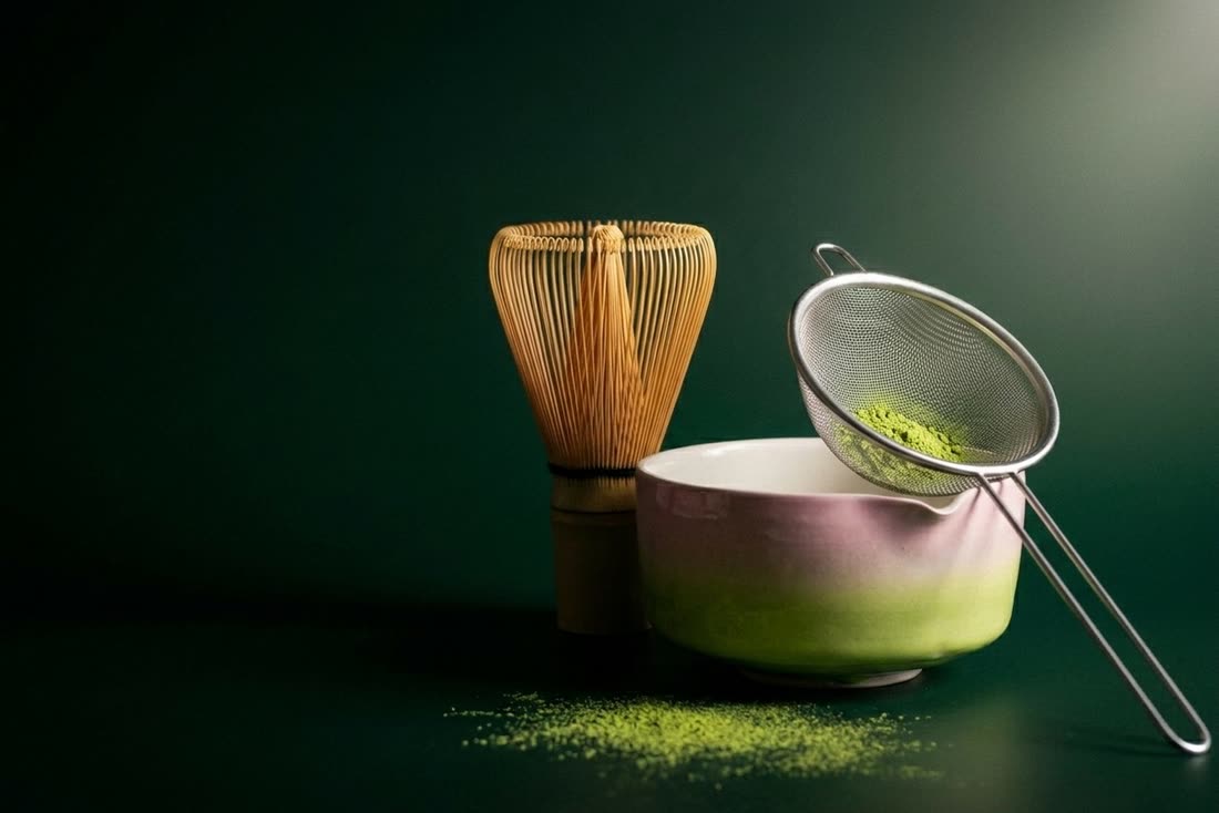 A bamboo whisk, a pink and green glazed matcha bowl and a steel sifter arranged together